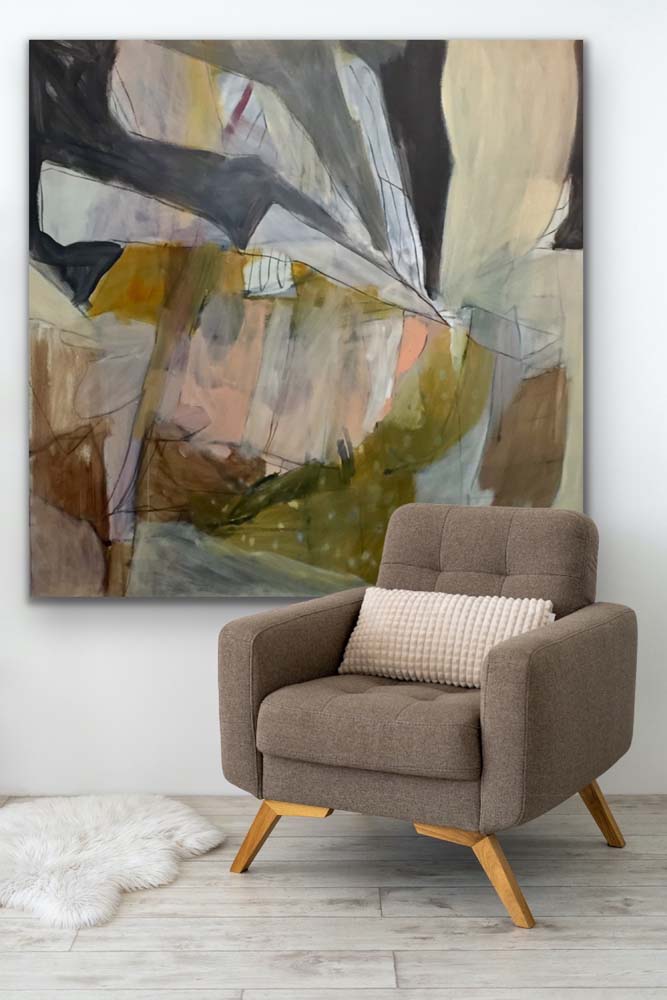abstract painting of irregular shapes in olive green, grey, brown and cream and mounted on a grey wall. There is a grey wooden floor below with a small white sheepskin rug and a brown chair with a cream cushion.