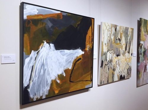 Three paintings hung on wall in line. The paintings are abstract forms in shades of cream and brown