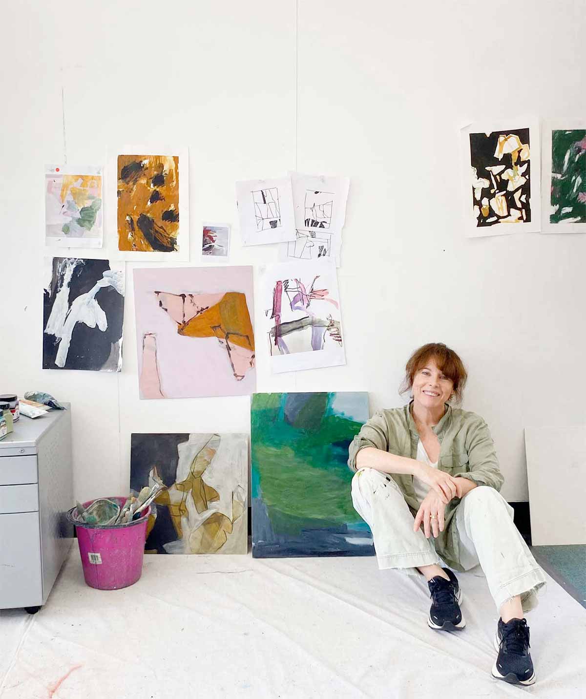 A smiling woman artist is sitting on a drop sheet on the floor with her back against a wall. She is surrounded by paintings and sketches. There is a pink bucket with artists tools by her side.